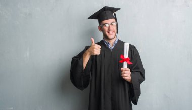 Beyaz delikanlı derecesi ile mutlu büyük holding lisansüstü üniforması gri grunge duvarın üzerinden gülümseme ok işareti, başparmak ile parmak, mükemmel işareti yapıyor