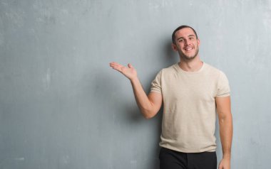 Neşeli sunulması ve kameraya bakarak avucunun ile işaret gülümseyen gri grunge duvarın üzerinden beyaz delikanlı.