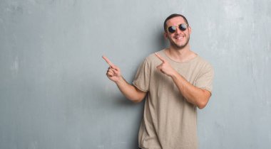 Gülümseyen ve iki elleri ve parmakları ile tarafına işaret kamera bakarak güneş gözlüğü takıyor gri grunge duvarın üzerinden beyaz delikanlı.