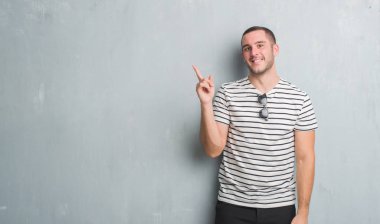 Kameraya bakarak yan el ve parmak ile işaret eden genç beyaz adam yüzünde kocaman bir gülümseme ile gri grunge duvarın üzerinden.