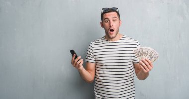 Dolar ve smarphone korkmuş şok bir sürpriz yüz, korkuyor ve heyecanlı korku ifadesi ile tutarak gri grunge duvarın üzerinden beyaz delikanlı
