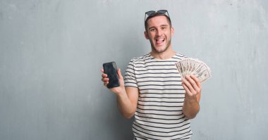 Beyaz delikanlı dolar ve smarphone dil yapışkanlık dışarı komik bir ifadeyle mutlu tutarak gri grunge duvarın üzerinden. Duygu kavramı.