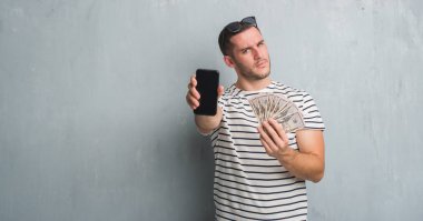 Beyaz delikanlı yüz çapraz silah ile dolar ve smarphone şüpheci ve sinir, onaylamayan ifade tutan gri grunge duvarın üzerinden. Negatif kişi.