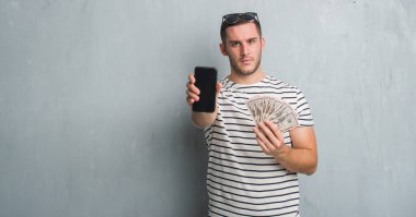 Beyaz delikanlı dolar ve smarphone ciddi ifade ile yüz tutan gri grunge duvarın üzerinden. Basit ve doğal kameraya bakıyor.