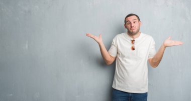 Beyaz genç güneş gözlüğü clueless ve şaşkın bir ifadeyle kollar ve eller kaldırdı giyen gri grunge duvarın üzerinden. Şüphe kavramı.