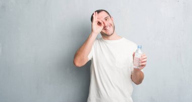 Gri grunge duvar holding şişe su ile mutlu yüz el parmaklarının arasından seyir göz üzerinde ok işaretiyle yapıyor gülümseyen Genç beyaz adam