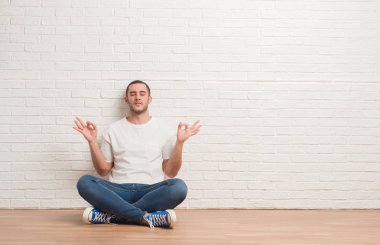 Beyaz tuğla duvarın üzerinden katta oturan genç beyaz adam sakin ol ve gülümseyen gözlerle kapalı parmak jestle meditasyon yapıyor. Yoga kavramı.