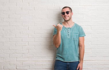 Genç beyaz adam işaret ve başparmak kadar mutlu yüz gülümseyen tarafı ile gösteren güneş gözlüğü takmış beyaz tuğla duvarın üzerinden ayakta