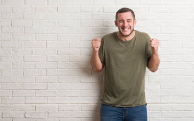Genç beyaz adam ayakta beyaz tuğla duvar silah ile başarı için heyecan üzerinde zafer kutluyor gülümseyen kaldırdı. Kazanan kavramı.