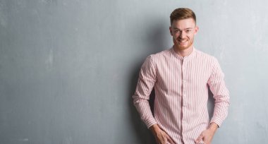 Genç Kızıl saçlı adam ayakta ve diş gösterilen kendine güvenen bir gülümsemeyle gülümseyerek mutlu bir yüz ile gri grunge duvarın üzerinden