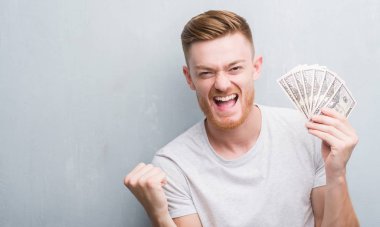 Duygu tezahürat genç Kızıl saçlı adam gurur çığlık ve zafer ve başarı çok heyecanlı, kutluyor dolar tutan gri grunge duvarın üzerinden