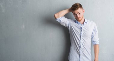 Young redhead business man over grey grunge wall confuse and wonder about question. Uncertain with doubt, thinking with hand on head. Pensive concept. clipart