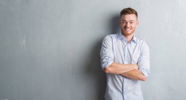 Gri grunge duvar mutlu yüz çapraz silah kameraya bakarak ile gülümseyen genç redhead iş adam. Olumlu bir kişi.