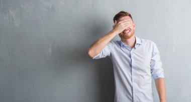 Genç redhead iş adamı gülümsüyor ve gözleri için sürpriz kapsayan yüz el ile gülüyor gri grunge duvarın üzerinden. Kör kavramı.