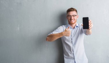 Gri grunge genç Kızıl saçlı adam akıllı telefon boş ekran mutlu ok işareti yapıyor büyük gülümseme ile gösterilen duvar, parmaklarıyla, mükemmel işaret parmak