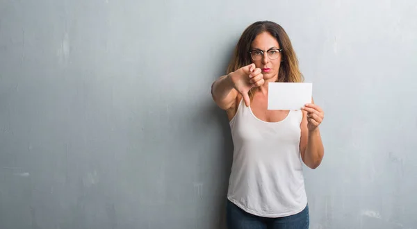 Middle age hispanic woman standing over grey grunge wall holding blank card with angry face, negative sign showing dislike with thumbs down, rejection concept