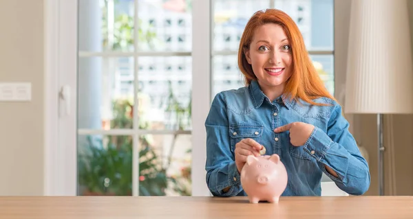 Redhead Woman Saves Money Piggy Bank Home Surprise Face Pointing — Stock Photo, Image