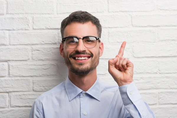 年轻的商业男子站在白色砖墙惊讶于一个想法或问题指向手指与快乐的脸 — 图库照片
