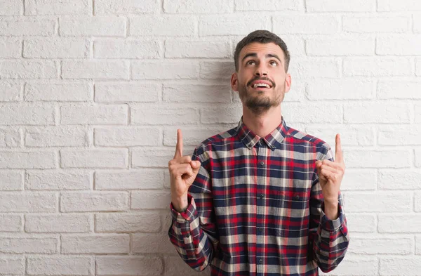 Genç yetişkin adam ayakta beyaz tuğla hayran ve sürpriz ve parmakları ile işaret duvar ve silah kaldırdı.