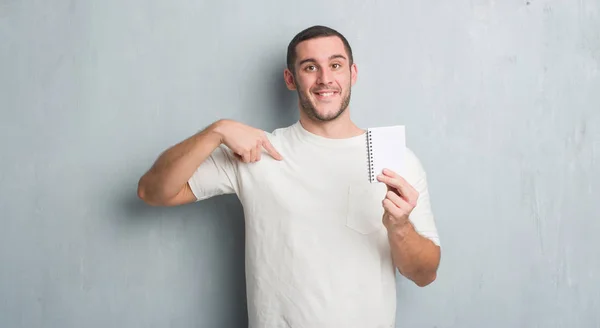 Kaukasische Jongeman Grijs Grunge Muur Weergegeven Notebook Met Verbazing Geconfronteerd — Stockfoto