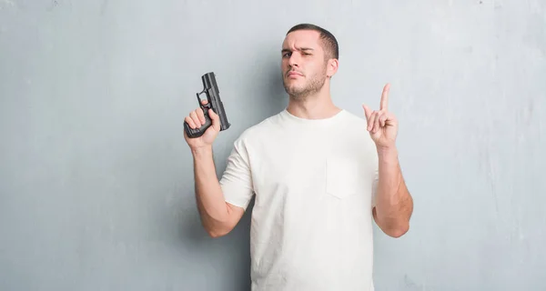 年轻的白种人犯罪男子在灰色的垃圾墙持有枪惊讶的想法或问题指向手指与快乐的脸 — 图库照片