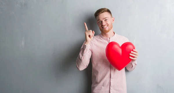 Young Redhead Man Grey Grunge Wall Holding Red Heart Surprised — Stock Photo, Image