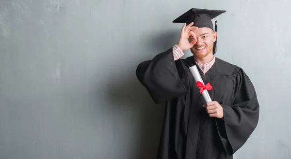 Young redhead man over grey grunge wall wearing graduate uniform holding degree with happy face smiling doing ok sign with hand on eye looking through fingers