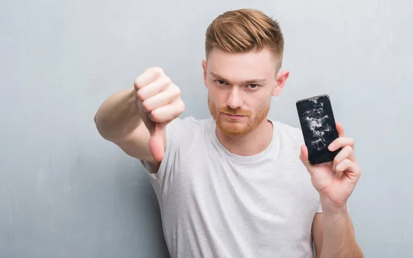 Joven Pelirrojo Sobre Pared Grunge Gris Sosteniendo Teléfono Inteligente Roto —  Fotos de Stock