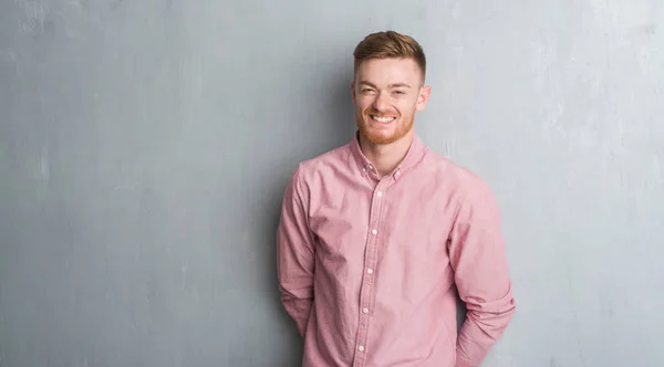 Young redhead man over grey grunge wall wearing pink shirt with a happy and cool smile on face. Lucky person.