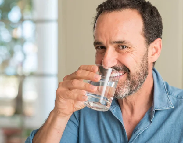 Middle age man drinking a glass of water with a happy face standing and smiling with a confident smile showing teeth