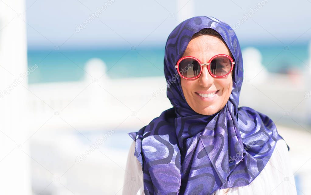 Middle age brunette arabian woman by the pool wearing sunglasses with a happy face standing and smiling with a confident smile showing teeth