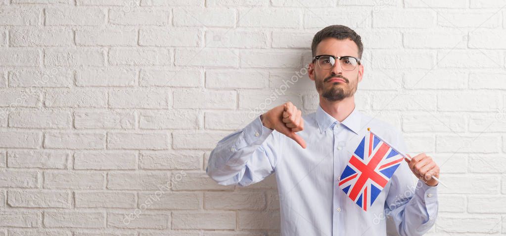 Young adult man over brick wall holding flag of United Kingdom with angry face, negative sign showing dislike with thumbs down, rejection concept