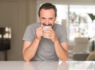 Orta yaş adam ayakta ve diş gösterilen kendine güvenen bir gülümsemeyle gülümseyerek mutlu bir yüz ile bir fincan kahve içme