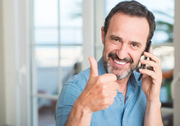 Middle age man using smartphone happy with big smile doing ok sign, thumb up with fingers, excellent sign