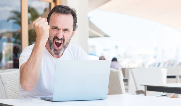 Handsome senior man using laptop at restaurant annoyed and frustrated shouting with anger, crazy and yelling with raised hand, anger concept