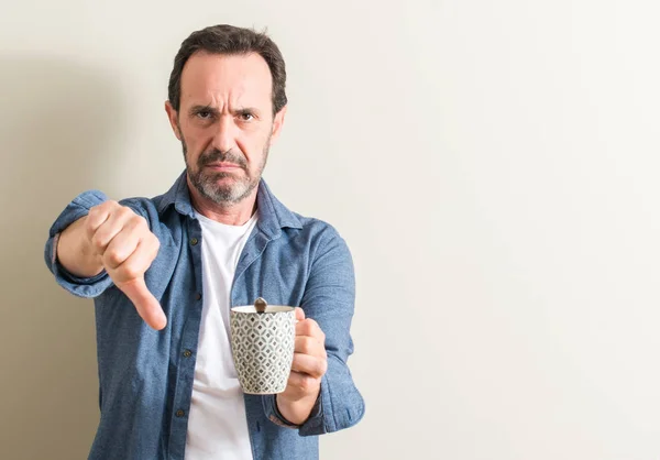 Senior man drinking coffee in a mug with angry face, negative sign showing dislike with thumbs down, rejection concept