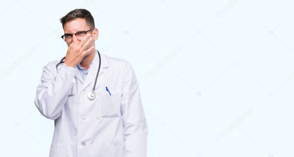 Handsome young doctor man smelling something stinky and disgusting, intolerable smell, holding breath with fingers on nose. Bad smells concept.