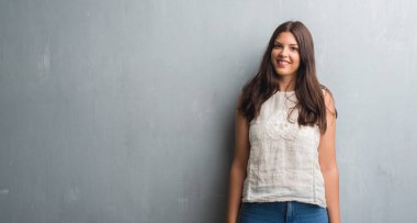 Grunge gri duvar ayakta ve diş gösterilen kendine güvenen bir gülümsemeyle gülümseyerek mutlu bir yüz ile genç esmer kadın