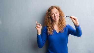 Gri grunge duvar optik gözlük çok mutlu el ve parmak ile tarafına işaret tutan genç Kızıl saçlı kadın