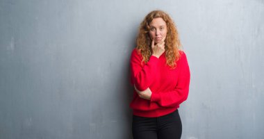 Genç redhead gri grunge duvar giyen üzerinde kırmızı kadın kazak emin çapraz kollar ve çeneme kaldırdı el ile gülümseme ile kameraya bakıyor. Pozitif düşünce.