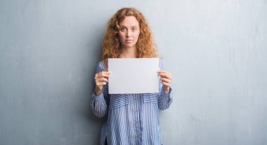 Gri grunge duvar holding boş kağıt sayfası acımak yüz ciddi düşünüyorum üstünde kendine güvenen bir ifade ile genç Kızıl saçlı kadın