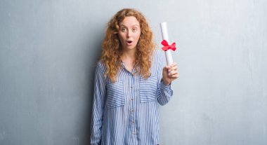 Genç redhead iş kadını diploma korkmuş şok bir sürpriz yüz, korkuyor ve heyecanlı korku ifadesi ile tutarak gri grunge duvarın üzerinden