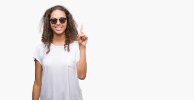 Gösterilen ve parmak numara ile yukarı dönük kendinden emin ve mutlu gülümseyen güneş gözlüğü takmış genç İspanyol kadın.