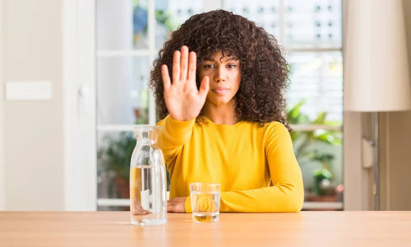 African american woman drinking a glass of water at home with open hand doing stop sign with serious and confident expression, defense gesture