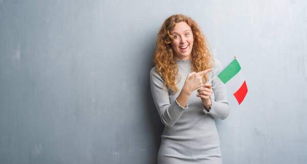 Young redhead woman over grey grunge wall holding flag of Italy very happy pointing with hand and finger