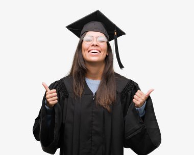 Mezun kap ve el ile pozitif jest yapıyor Tekdüzen başarı işareti giyen genç İspanyol kadın başparmak gülümseyen ve mutlu. Neşeli bir ifadeyle, kazanan jest kameraya bakıyor.