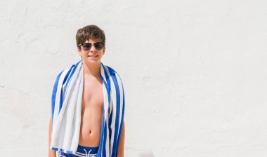 Tatil günlerinde ayakta ve diş gösterilen kendine güvenen bir gülümsemeyle gülümseyerek mutlu bir yüz ile bir donanma havlu giyen genç çocuk