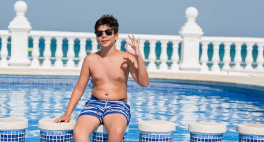 Parmak, mükemmel sembolü ile ok işareti yaparken plajda yüzme havuzunda genç çocuk tatilde