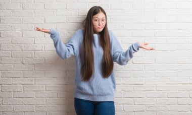 Genç Chinise kadın beyaz tuğla duvar clueless ve şaşkın bir ifadeyle kollar ve eller kaldırdı. Şüphe kavramı.