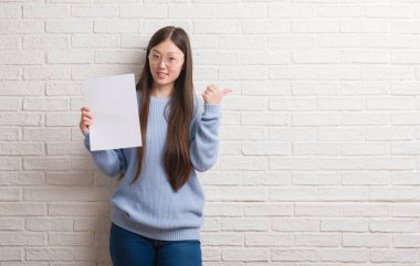 Genç Çinli kadın işaret ve başparmak kadar mutlu yüz gülümseyen tarafı ile gösterilen boş kağıt tutan tuğla duvarın üzerinden